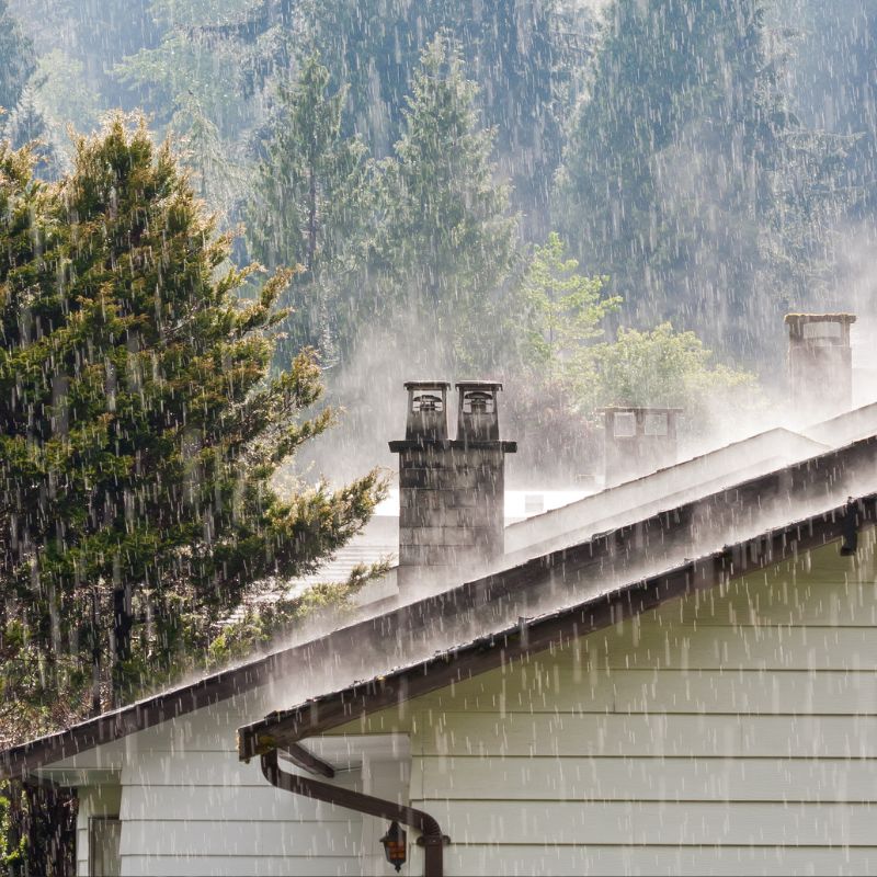 rain falling on a home with a chimney with trees in the background