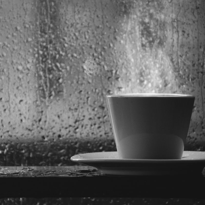 a white cup with steam coming out of the top on a white plate in front of a window that shows it's raining outside